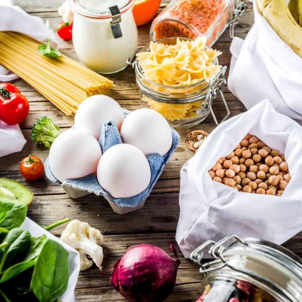 Healthy cheap budget friendly foods, like gluten-free pasta, eggs, beans, broccoli, tomatoes, and lentils on a wooden kitchen table.